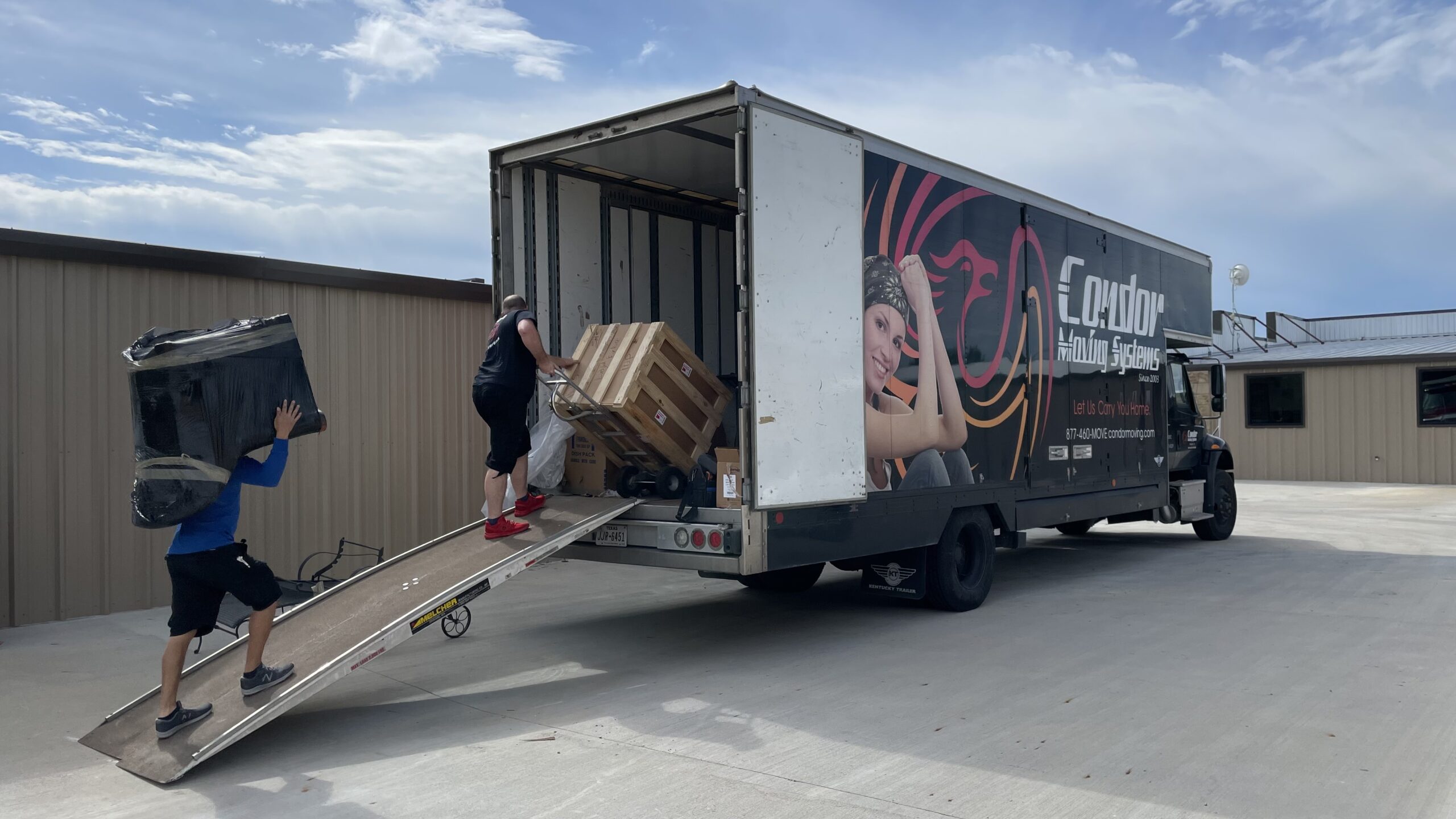 Condor movers carrying a wrapped television and a wooden crate up the ramp into a moving truck